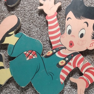 Peut inclure: Puzzle en bois vintage repr&eacute;sentant un gar&ccedil;on de dessin anim&eacute; en salopette turquoise et une chemise ray&eacute;e rouge et blanche. Le gar&ccedil;on a les cheveux noirs et les joues roses, un bras lev&eacute;. Les pi&egrave;ces du puzzle sont sur un tapis gris.