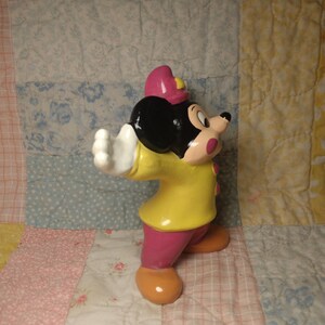 Puede incluir: Una figurita de cer&aacute;mica de Mickey Mouse vestido de bombero. Lleva una chaqueta amarilla, pantalones rosas y un gorro rojo. Tiene orejas negras y una nariz negra.