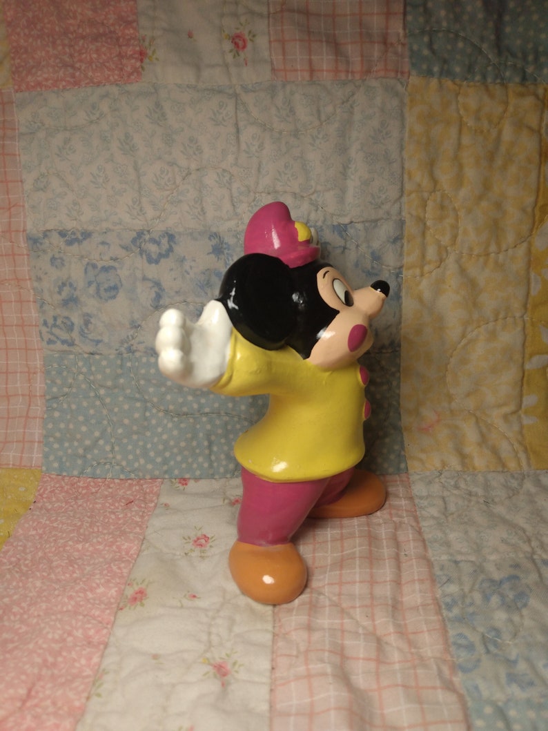Puede incluir: Una figurita de cer&aacute;mica de Mickey Mouse vestido de bombero. Lleva una chaqueta amarilla, pantalones rosas y un gorro rojo. Tiene orejas negras y una nariz negra.