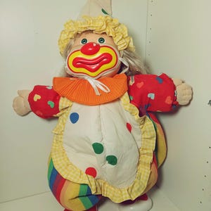 May include: A clown doll with a red nose and a big smile, wearing a colorful outfit with a yellow and white checkered pattern, a rainbow striped skirt, and a yellow and white hat with a red pom-pom.