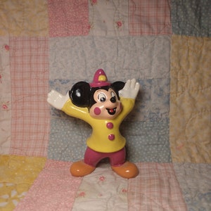 Puede incluir: Una figurita de cer&aacute;mica de Mickey Mouse vestido de bombero. Lleva una chaqueta amarilla con botones rojos, un sombrero rosa y pantalones negros. Est&aacute; de pie con los brazos en alto y una sonrisa en la cara.