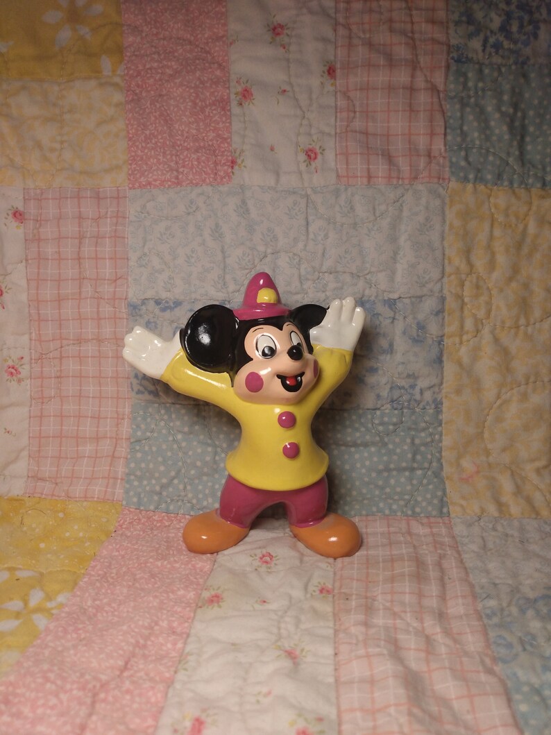 Puede incluir: Una figurita de cer&aacute;mica de Mickey Mouse vestido de bombero. Lleva una chaqueta amarilla con botones rojos, un sombrero rosa y pantalones negros. Est&aacute; de pie con los brazos en alto y una sonrisa en la cara.
