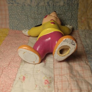 Puede incluir: Una figurita de cer&aacute;mica amarilla, rosa y negra de Mickey Mouse. La figurita est&aacute; tumbada de espaldas con los brazos extendidos. La figurita tiene un agujero en la parte inferior, lo que sugiere que podr&iacute;a ser un salero o un pimentero.