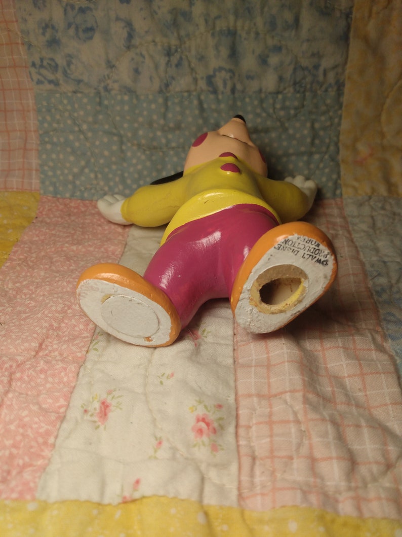 Puede incluir: Una figurita de cer&aacute;mica amarilla, rosa y negra de Mickey Mouse. La figurita est&aacute; tumbada de espaldas con los brazos extendidos. La figurita tiene un agujero en la parte inferior, lo que sugiere que podr&iacute;a ser un salero o un pimentero.