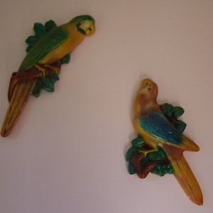 May include: Two ceramic parrot figurines with green, blue, and yellow paint. The parrots are perched on branches with green leaves.
