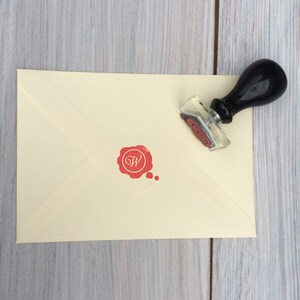 May include: A cream envelope with a red wax seal with the letter "W" inside. A black and silver stamp with a handle is next to the envelope.
