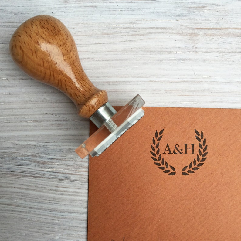May include: A wooden stamp with a metal base and a handle. The stamp is used to create a design with a laurel wreath and the initials "A&H".