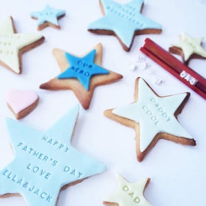 Puede incluir: Surtido de galletas en forma de estrella con glaseado azul, amarillo y rosa. Algunas galletas tienen texto que dice "Happy Father's Day Love Ella & Jack", "Our Hero" y "Daddy Cool". Tambi&eacute;n hay una herramienta roja con la palabra "DAD".