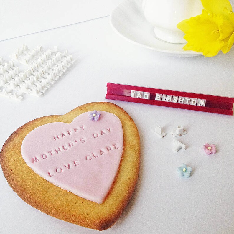 Puede incluir: Una galleta en forma de coraz&oacute;n con un coraz&oacute;n de fondant rosa que dice "Happy Mother's Day Love Clare". La galleta est&aacute; decorada con peque&ntilde;as chispas en forma de flor. Un soporte de pl&aacute;stico rojo muestra el texto "MOTHER'S DAY".