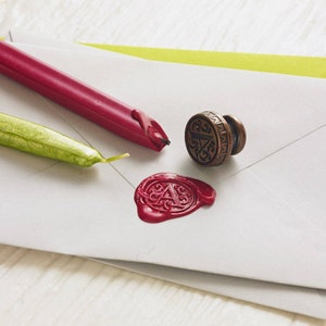 May include: A red wax seal stamp with a decorative design and a red wax seal with the letter A in the centre. The stamp is made of metal and has a handle. The wax seal is on a white envelope.