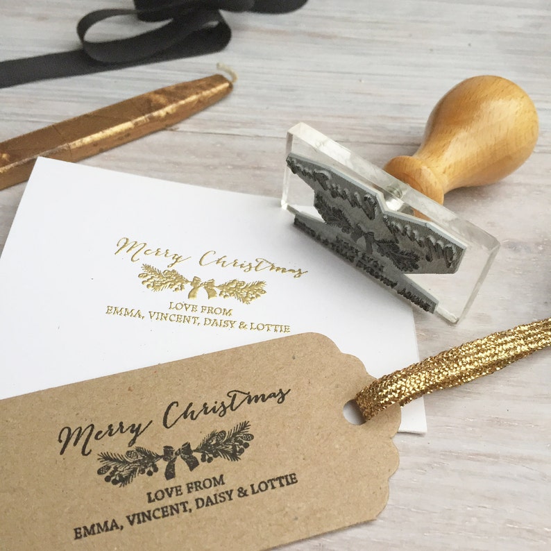 May include: A clear acrylic stamp with a gold foil design of a Christmas wreath with a bow. The stamp reads "Merry Christmas" and "Love from Emma, Vincent, Daisy & Lottie".