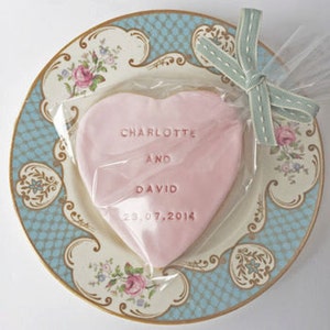 Puede incluir: Una galleta rosa en forma de coraz&oacute;n con los nombres "Charlotte y David" y la fecha "23.07.2014" grabados, envuelta en celof&aacute;n transparente y atada con una cinta azul claro. La galleta descansa sobre un plato decorativo con un dise&ntilde;o azul y floral.