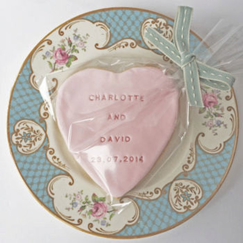 Puede incluir: Una galleta rosa en forma de coraz&oacute;n con los nombres "Charlotte y David" y la fecha "23.07.2014" grabados, envuelta en celof&aacute;n transparente y atada con una cinta azul claro. La galleta descansa sobre un plato decorativo con un dise&ntilde;o azul y floral.