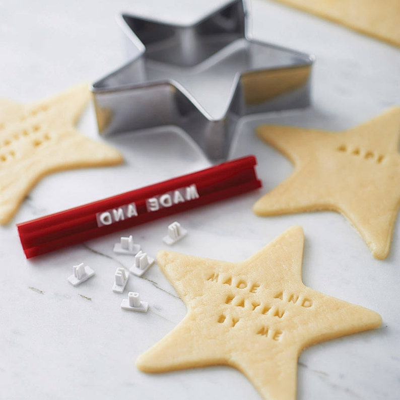 Puede incluir: Un cortador de galletas en forma de estrella y galletas con el texto "MADE AND EATEN BY ME" estampado. Tambi&eacute;n se ve una herramienta de estampado roja y sellos de letras individuales. Las galletas son de color marr&oacute;n claro.