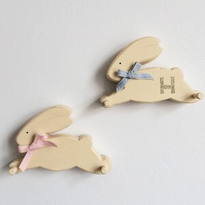 May include: Two wooden bunny shaped hooks with a light yellow finish. The bunny on the left has a pink bow and the bunny on the right has a blue bow. The bunny on the right has the letter "H" written on it in gold glitter.