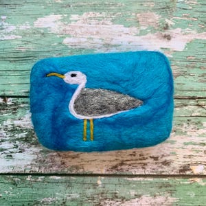 May include: A teal blue felted soap bar with a white seagull design. The seagull has yellow legs and a black eye.