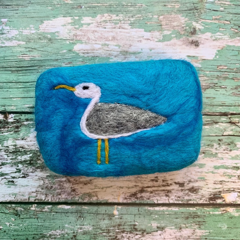 May include: A teal blue felted soap bar with a white seagull design. The seagull has yellow legs and a black eye.