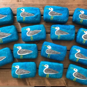 May include: A collection of 14 teal blue felted soap bars with a white seagull design. Each bar features a seagull with yellow feet and a white body on a teal blue background.