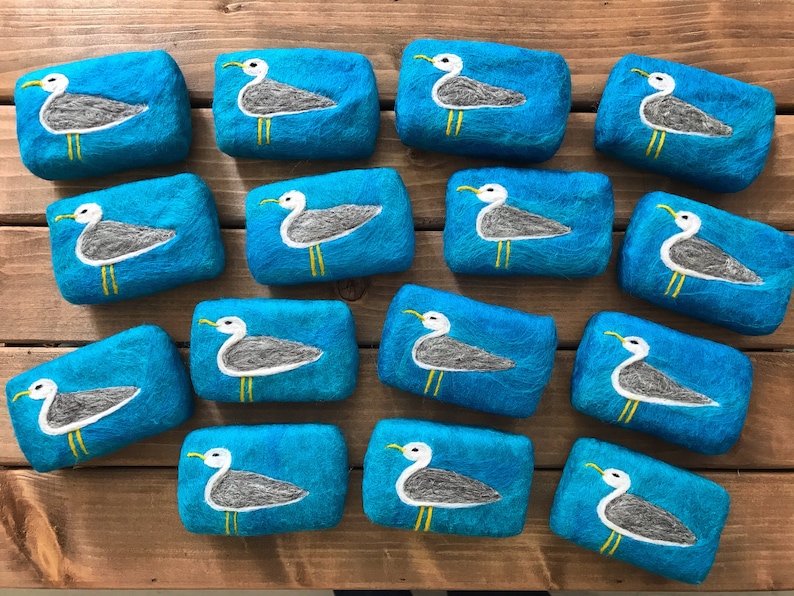 May include: A collection of 14 teal blue felted soap bars with a white seagull design. Each bar features a seagull with yellow feet and a white body on a teal blue background.