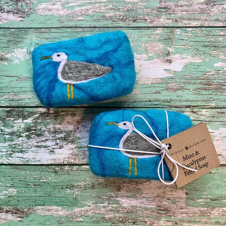 May include: Two teal blue felted soap bars with a white seagull design. The soap bars are tied with white twine and a brown tag that reads "Herbal Harmony Mint & Eucalyptus Felted Soap".