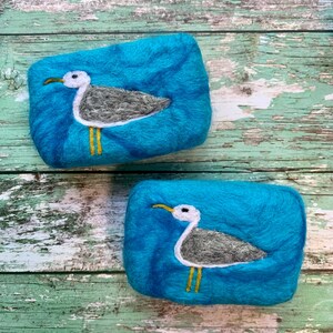 May include: Two blue felted soap bars with a white and gray seagull design. The seagulls have yellow beaks and legs.