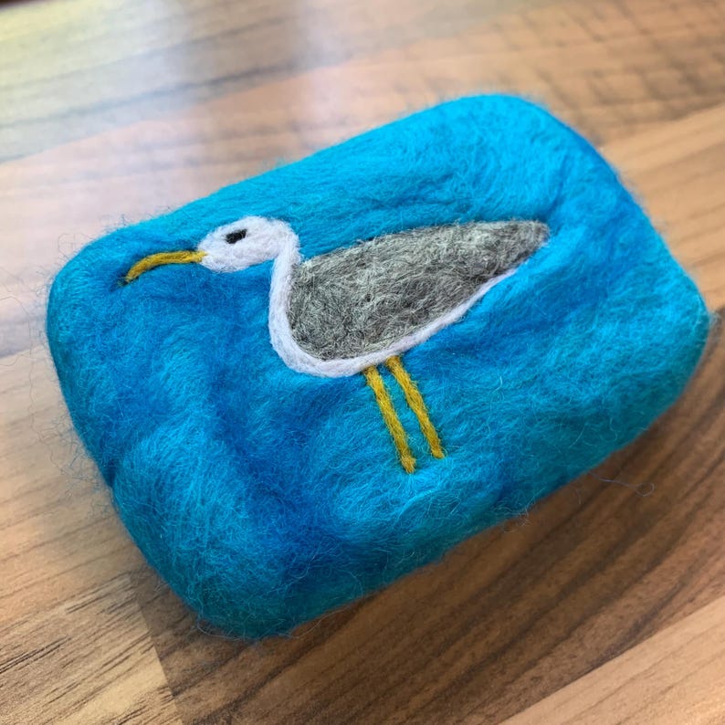 May include: A teal blue felted soap dish with a white and gray seagull design. The seagull has yellow legs and a black beak.