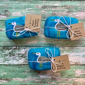 May include: Three bars of felted soap wrapped in twine and decorated with a seagull design. The soap bars are turquoise blue with a white seagull and yellow feet. The labels on the soap bars read "Mint & Eucalyptus Felted Soap" and "Mint & Bergamot Felted Soap".