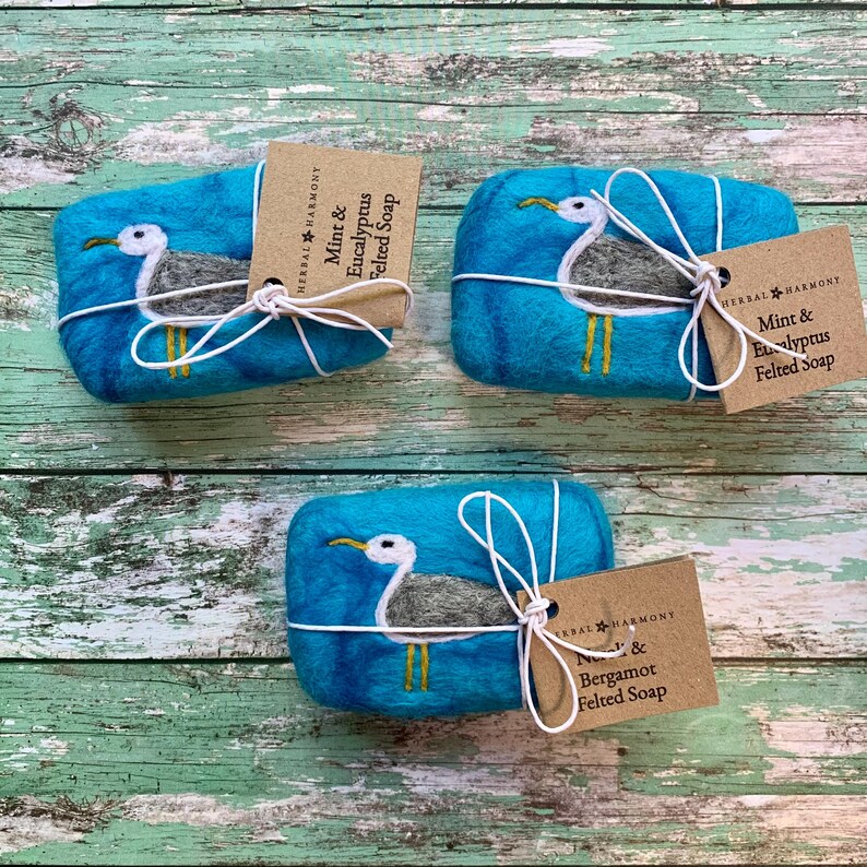 May include: Three bars of felted soap wrapped in twine and decorated with a seagull design. The soap bars are turquoise blue with a white seagull and yellow feet. The labels on the soap bars read "Mint & Eucalyptus Felted Soap" and "Mint & Bergamot Felted Soap".