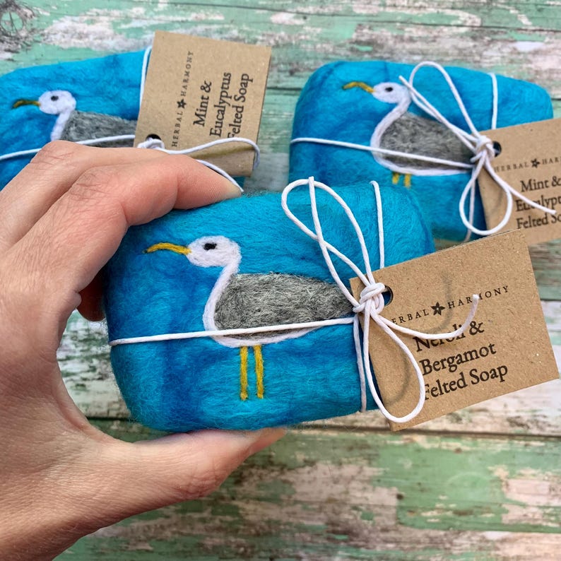May include: Three handmade felted soap bars wrapped in twine with a tag. Each bar features a seagull design and is tied with a white ribbon. The tags read "Mint & Eucalyptus Felted Soap", "Neroli & Bergamot Felted Soap", and "Mint & Eucalyptus Felted Soap".