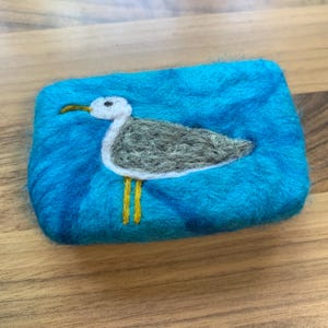 May include: A blue felted soap dish with a white and gray seagull design. The seagull has yellow legs and a white beak.
