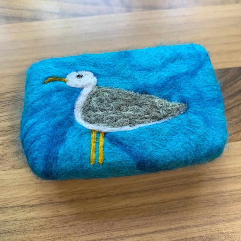 May include: A blue felted soap dish with a white and gray seagull design. The seagull has yellow legs and a white beak.