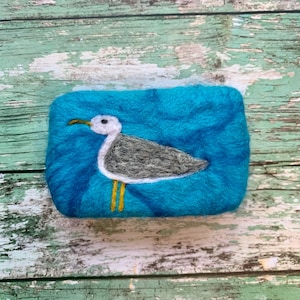 May include: A teal blue felted soap with a white and gray seagull design. The seagull has yellow feet.