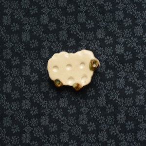 May include: A white ceramic sheep figurine with brown accents. The sheep has a rounded body and a small head with a brown nose. The sheep is sitting on a dark blue fabric with a floral pattern.