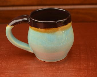 Può includere: Una tazza in ceramica fatta a mano con un corpo arrotondato e un manico curvo. La tazza presenta una sfumatura di colori, tra cui un bordo marrone scuro, una fascia dorata e un corpo azzurro. La tazza è progettata per bevande calde.