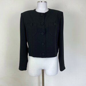 May include: A black cropped jacket with long sleeves and a round neckline. The jacket has large, round buttons down the front and on the pockets. The jacket is displayed on a white mannequin.