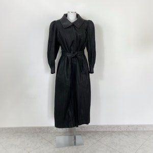 Peut inclure: Un trench-coat en cuir noir avec une ceinture à la taille et un col. Le manteau a une manche légèrement bouffante et une silhouette longue et fluide.