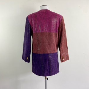 May include: A colour-blocked jacket in shades of purple, burgundy, and magenta. The jacket has a textured, snakeskin-like pattern and gold buttons on the sleeves. The garment is displayed on a mannequin.