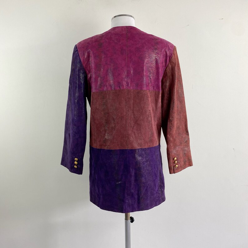 May include: A colour-blocked jacket in shades of purple, burgundy, and magenta. The jacket has a textured, snakeskin-like pattern and gold buttons on the sleeves. The garment is displayed on a mannequin.