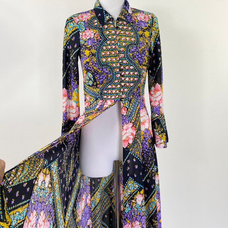 Puede incluir: Un vestido largo de manga larga con fondo negro y estampado floral colorido. El vestido tiene cuello alto, cierre de botones en la parte delantera y una larga abertura en la parte delantera. Las mangas son largas y acampanadas.
