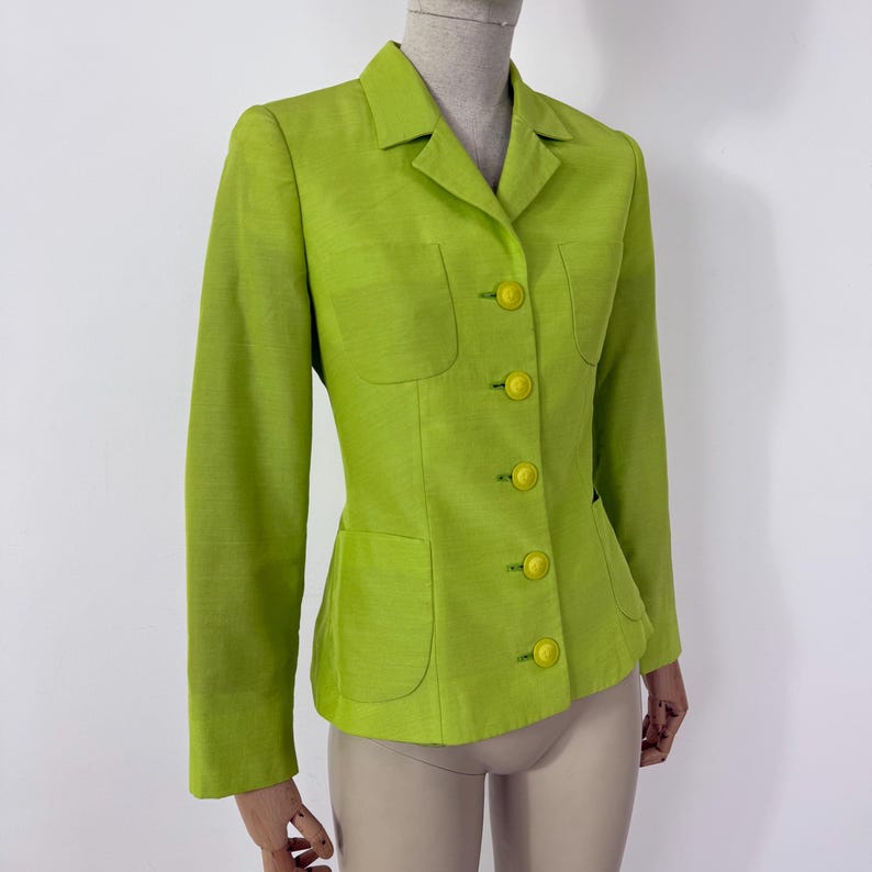 May include: A lime green blazer with a tailored fit. The blazer features a notched collar, two front pockets, and bright yellow buttons. The long-sleeved jacket is displayed on a mannequin.