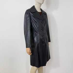 May include: A black leather coat with a collared neckline and button-down closure. The coat features long sleeves, a tailored fit, and a knee-length hemline. The coat has a textured appearance and a classic design.