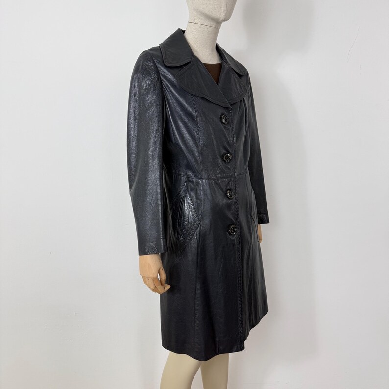 May include: A black leather coat with a collared neckline and button-down closure. The coat features long sleeves, a tailored fit, and a knee-length hemline. The coat has a textured appearance and a classic design.
