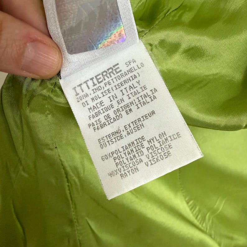 May include: A white label with black text, featuring the brand name "ITTIERRE" and manufacturing details. The label is held against a bright green fabric, with the text indicating the item is made in Italy.