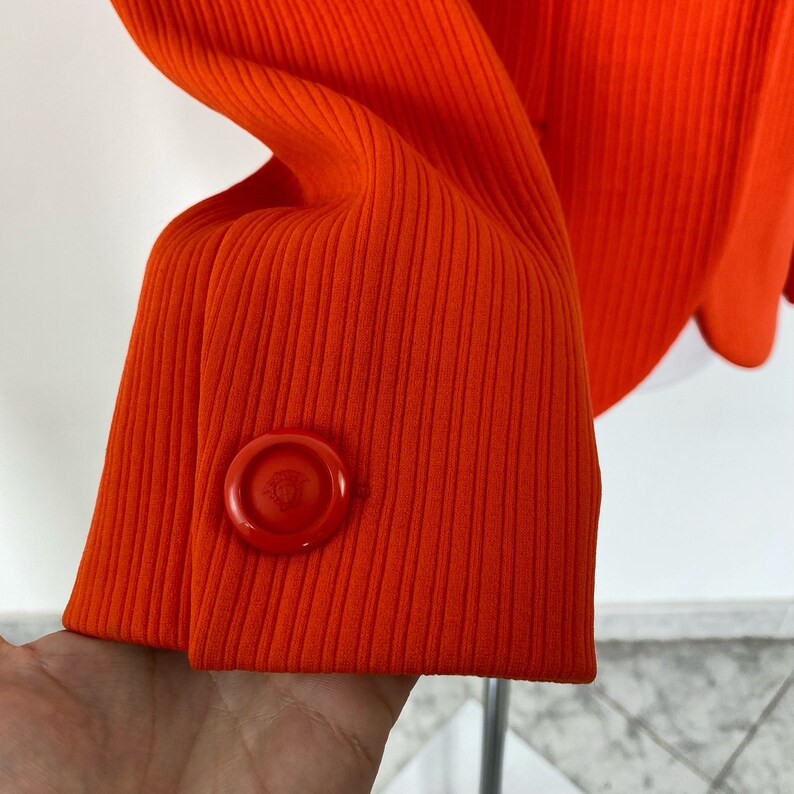 May include: Orange ribbed blazer with a single button closure on the sleeve. The button is red and has a logo on it.