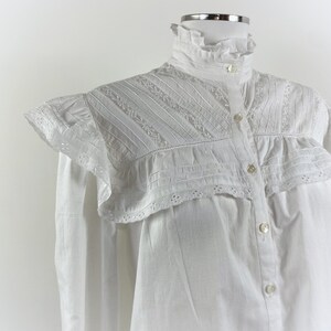 Puede incluir: Una blusa blanca con cuello alto con volantes y cierre de botones. Los hombros presentan un dise&ntilde;o decorativo en capas con detalles bordados y plisados. Las mangas largas son visibles en los laterales.