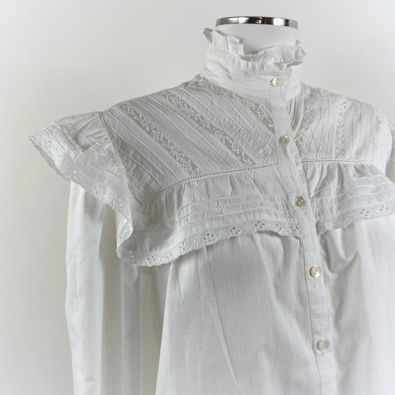 Puede incluir: Una blusa blanca con cuello alto con volantes y cierre de botones. Los hombros presentan un dise&ntilde;o decorativo en capas con detalles bordados y plisados. Las mangas largas son visibles en los laterales.