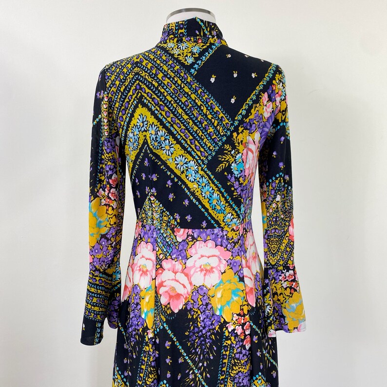 Puede incluir: Un vestido de manga larga con fondo negro y estampado floral colorido. El vestido tiene cuello alto y mangas acampanadas.