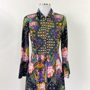 Puede incluir: Un vestido largo de manga larga, negro, con estampado floral en tonos rosa, morado, amarillo y azul. El vestido tiene un frente abotonado y un cuello.