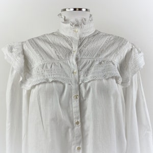 Puede incluir: Una camisa blanca abotonada con cuello alto con volantes y detalles de hombros con volantes. La camisa presenta intrincados encajes y pliegues en el canes&uacute; y las mangas, y se abrocha en la parte delantera con peque&ntilde;os botones redondos.