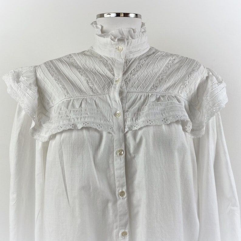 Puede incluir: Una camisa blanca abotonada con cuello alto con volantes y detalles de hombros con volantes. La camisa presenta intrincados encajes y pliegues en el canes&uacute; y las mangas, y se abrocha en la parte delantera con peque&ntilde;os botones redondos.